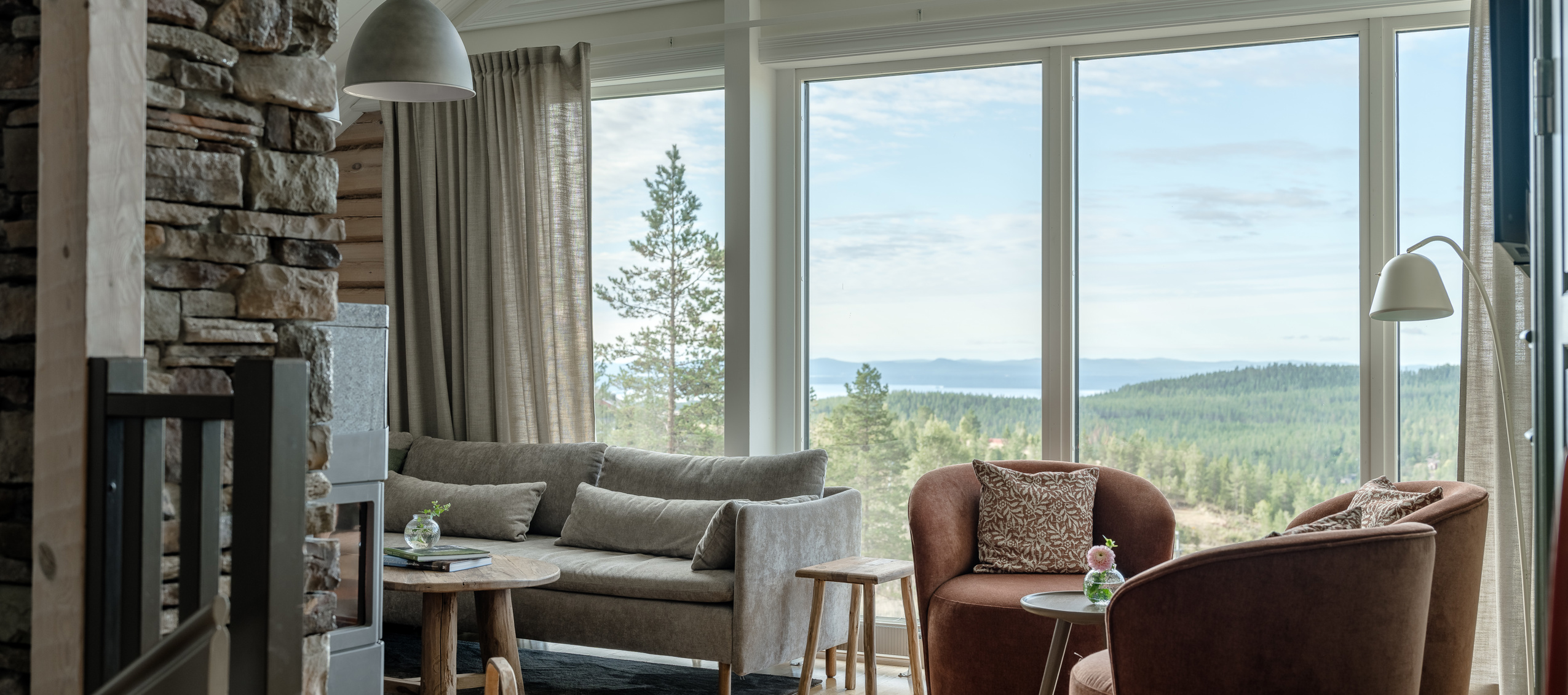 Ett ljust vardagsrum med stora panoramafönster som vetter mot skog och berg, möblerat med soffor och fåtöljer.