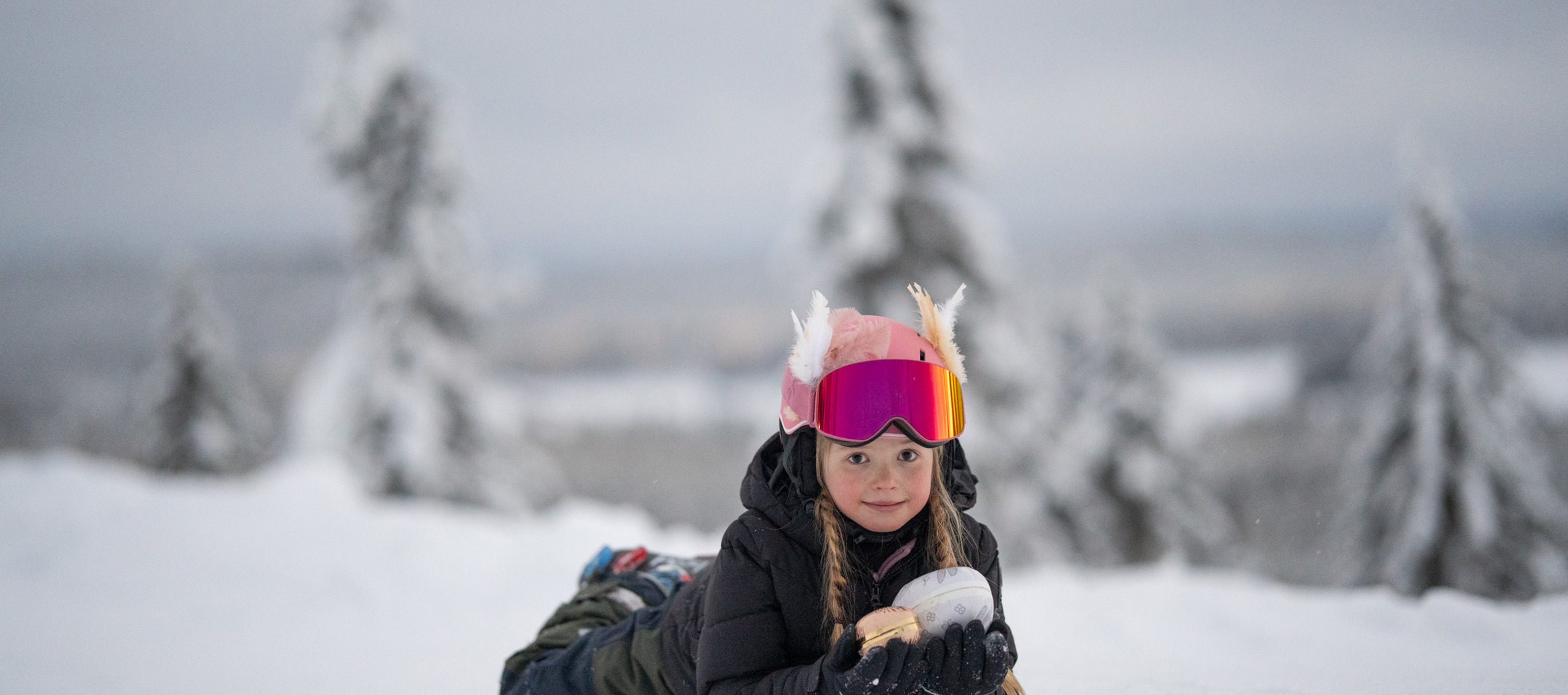 Barn ligger i snön med skidglasögon och håller i en snöboll, med snötäckt skog i bakgrunden.