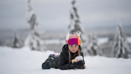 Barn ligger i snön med skidglasögon och håller i en snöboll, med snötäckt skog i bakgrunden.