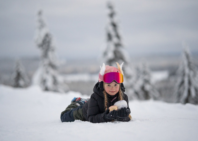 Barn ligger i snön med skidglasögon och håller i en snöboll, med snötäckt skog i bakgrunden.