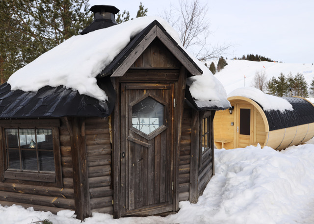 A grill hut and sauna surrounded by snow in a winter landscape, with ski slopes in the background.