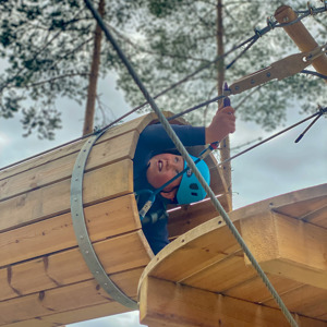 Barn med blå hjälm klättrar genom ett hinder i en höghöjdsbana i skogen, säkrad med sele och karbinhake.