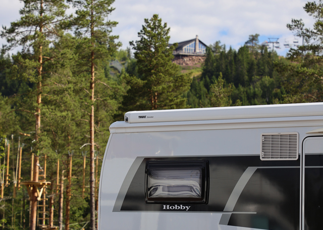 Husbil parkerad vid skogsbryn med träd i bakgrunden och en stuga uppe på höjden i fjärran.