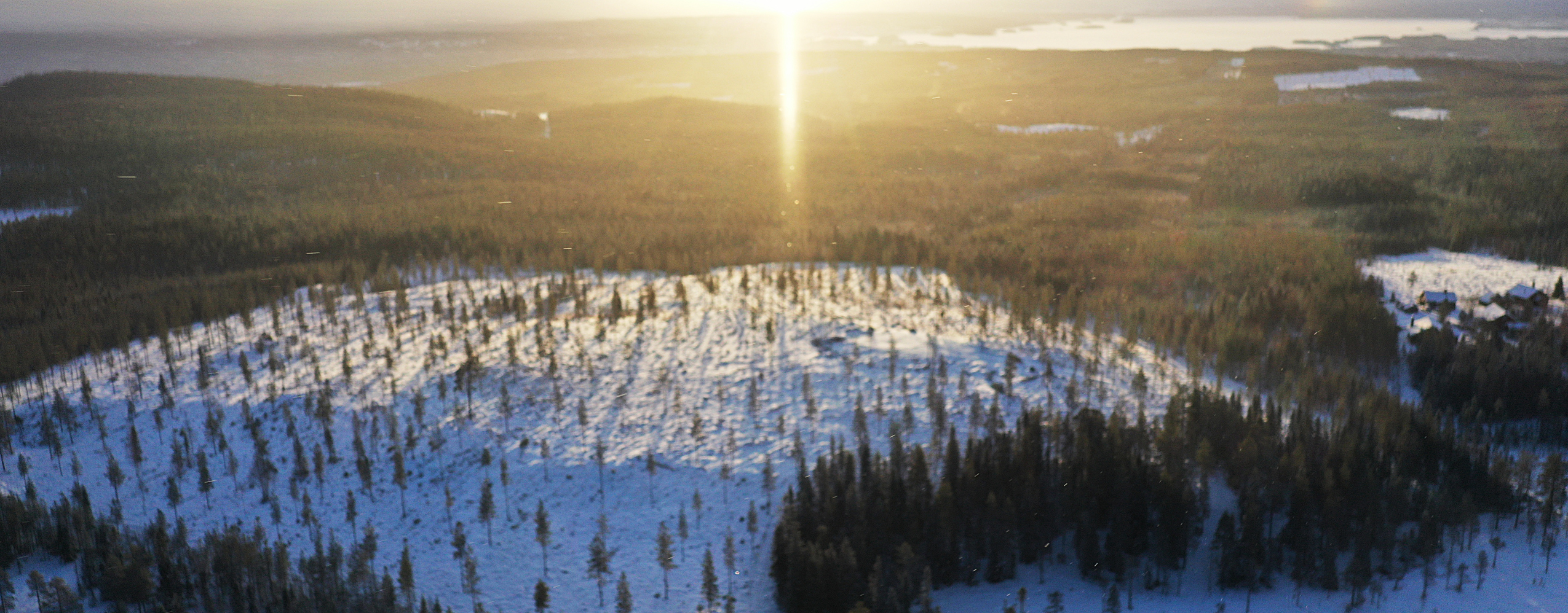 Flygfoto över ett snötäckt skogsområde i soluppgång, med solstrålar som bryter genom horisonten och visar vyer över fjäll, skog och sjöar.