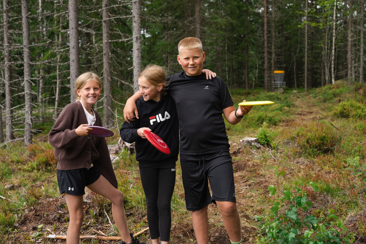 Tre barn står med discar i händerna vid en discgolfbana i skogen och ler mot kameran.