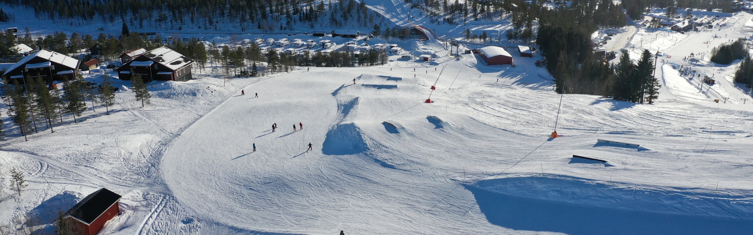 Luftfoto af en snowpark med skiløbere, hop og rails, omgivet af hytter og skov i vinterlandskab