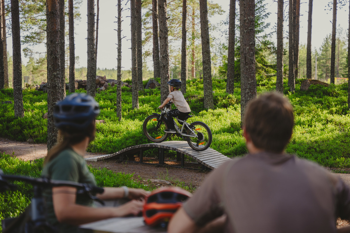 Ett barn cyklar på en teknikbana i skogen medan två vuxna tittar på från sidan.