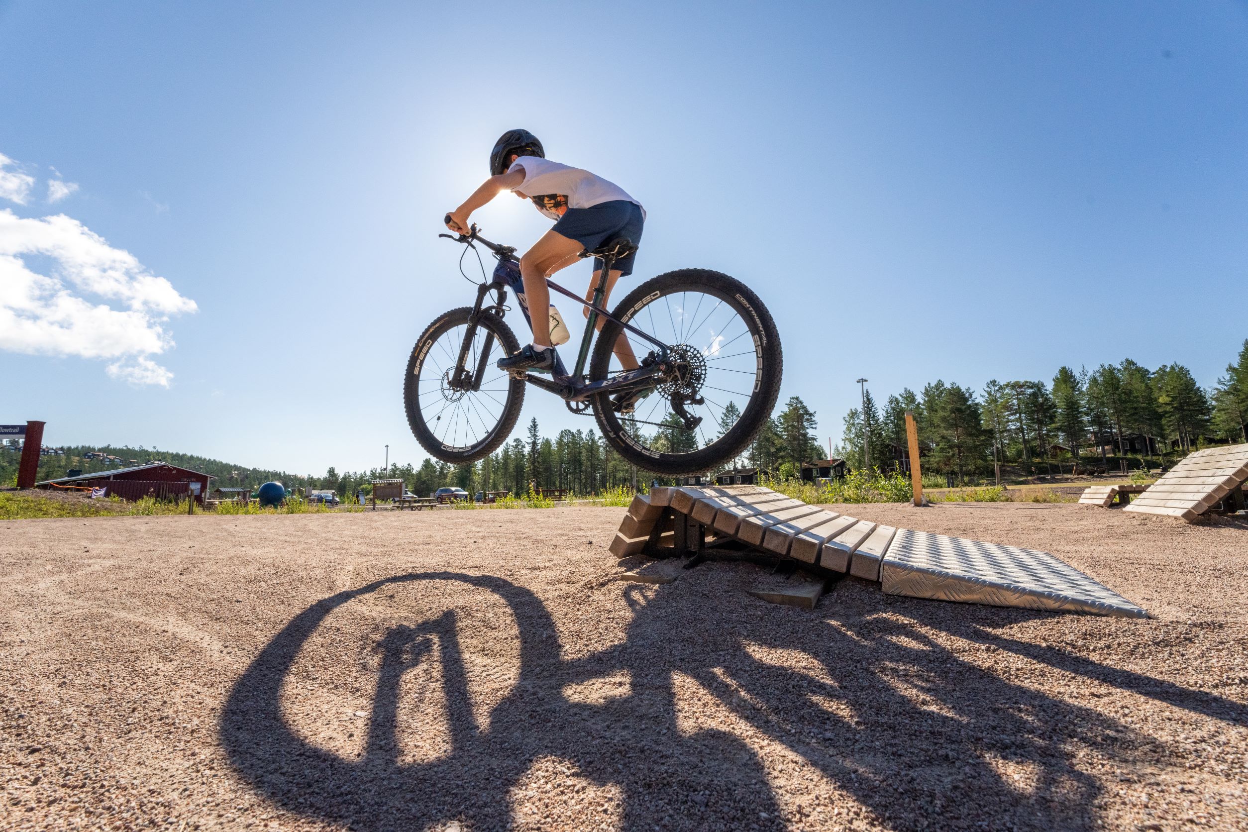A person jumps with a mountain bike off a ramp in a bike park, their shadow clearly cast on the ground below. Sunny weather and blue sky, with forest and buildings in the background.