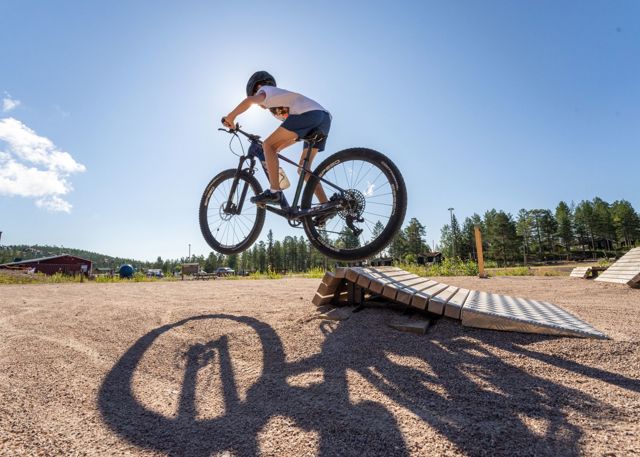 En person hoppar med mountainbike från ett hopp i en cykelpark, med sin skugga tydligt projicerad på marken under. Soligt väder och blå himmel, skog och byggnader syns i bakgrunden.