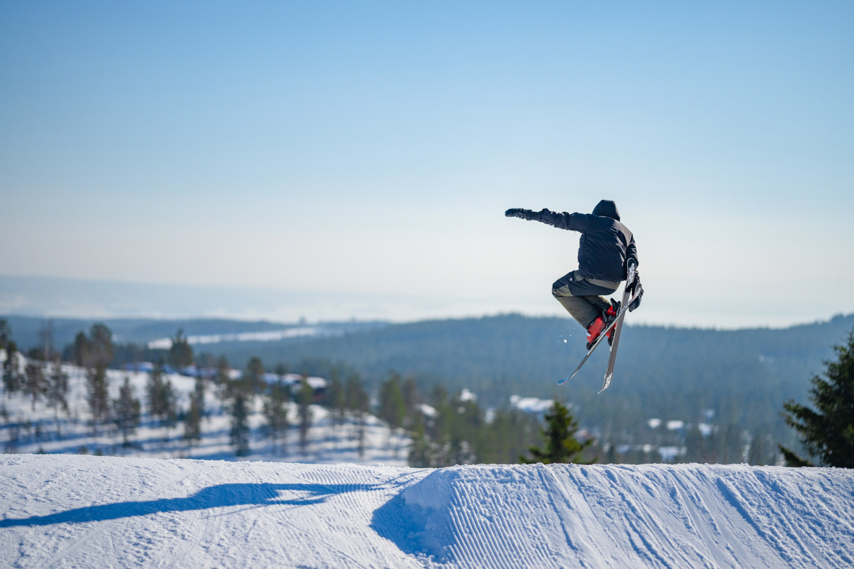 Skiløber laver et hop i en snowpark med snelandskab og skov i baggrunden.