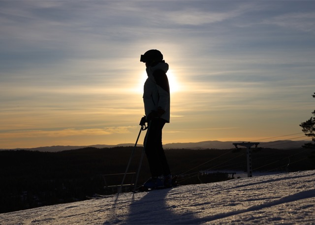Silhuett av en skidåkare som står stilla i backen i motljus från en dramatisk solnedgång över fjällandskapet.
