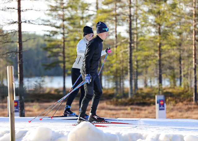 Two people cross-country skiing on a snowy trail through the forest, with a lake and evergreen trees in the background.