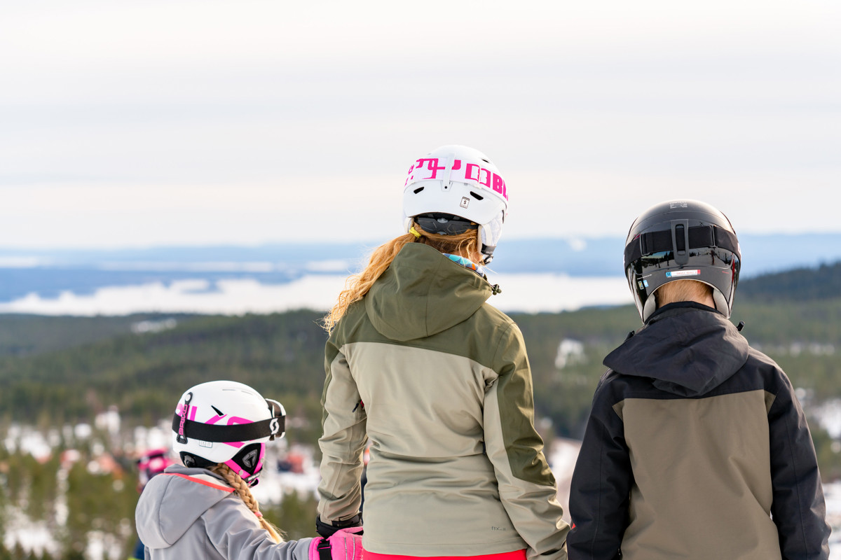 An adult and two children wearing ski helmets look out over the snowy landscape from the top of a ski slope at Orsa Grönklitt.