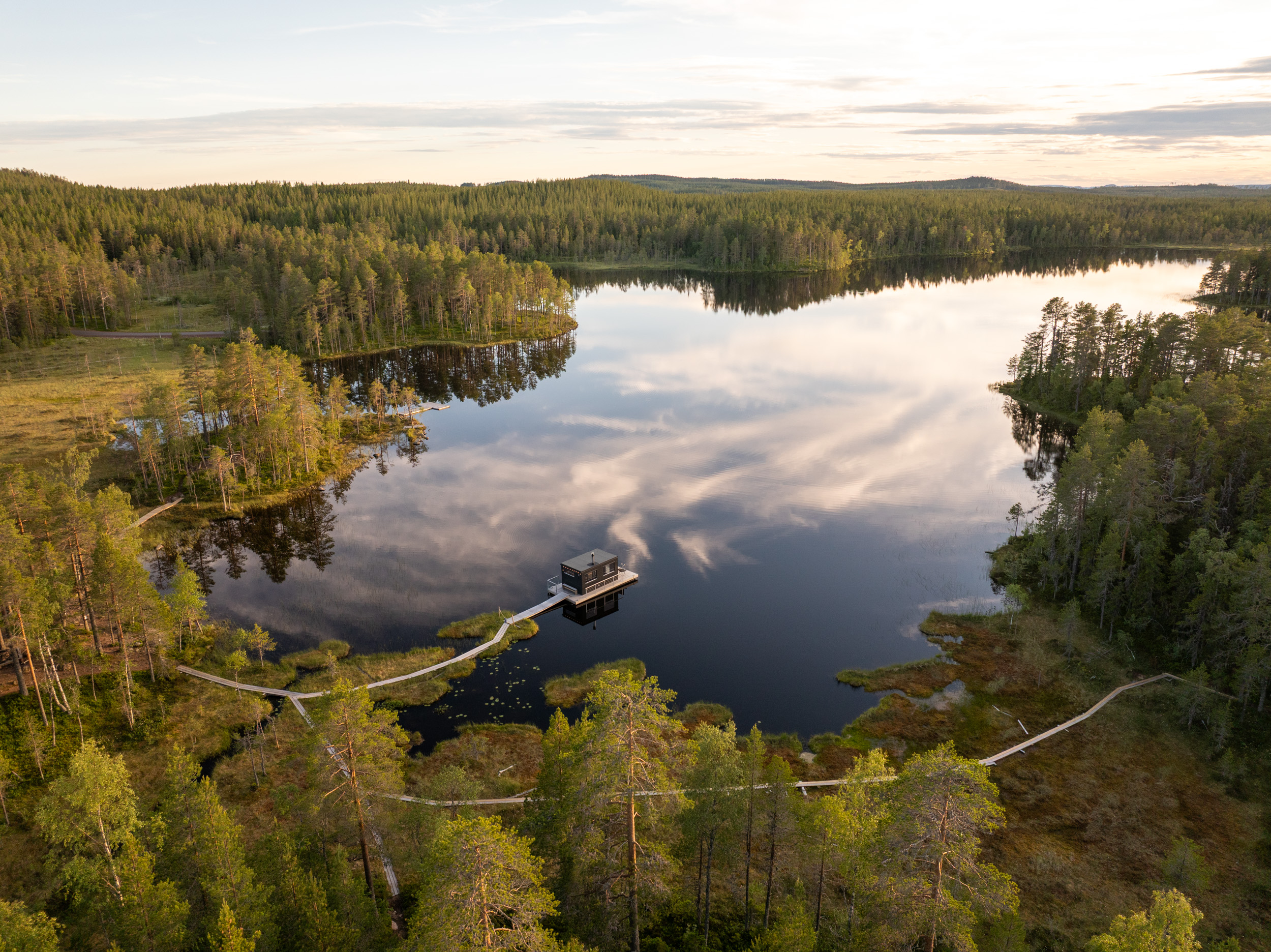 Flygbild över sjö omgiven av skog med gångbro och liten byggnad på vattnet.