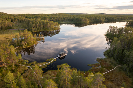 Aerial view of a lake surrounded by forest with a walkway and a small building on the water.