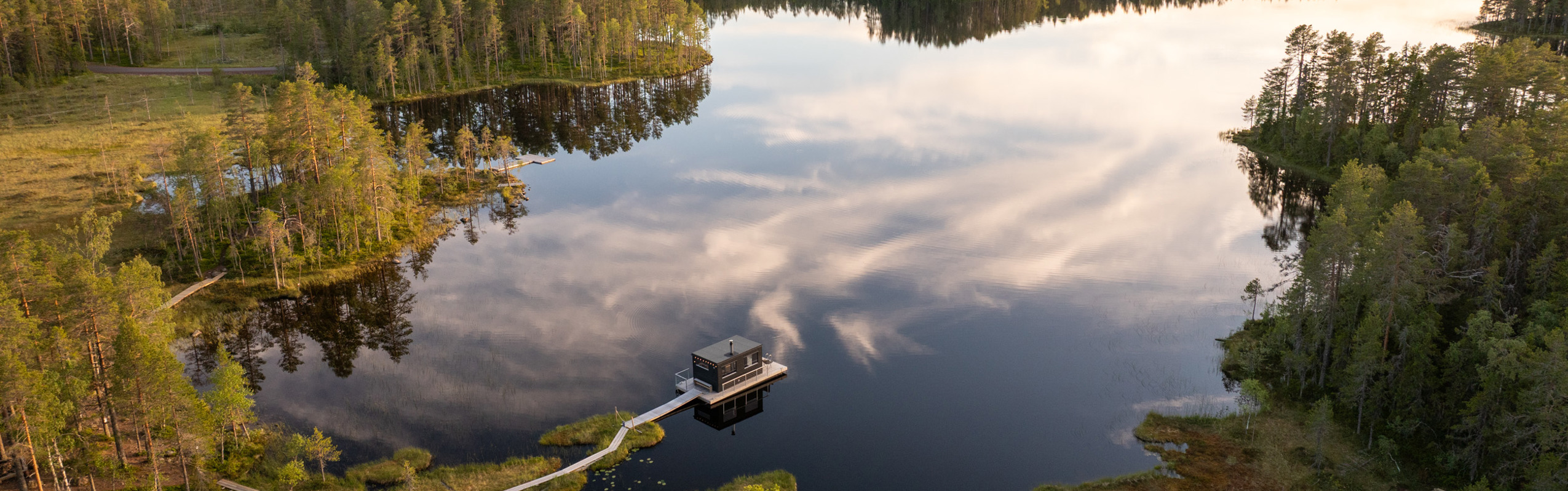 Aerial view of a lake surrounded by forest with a walkway and a small building on the water.
