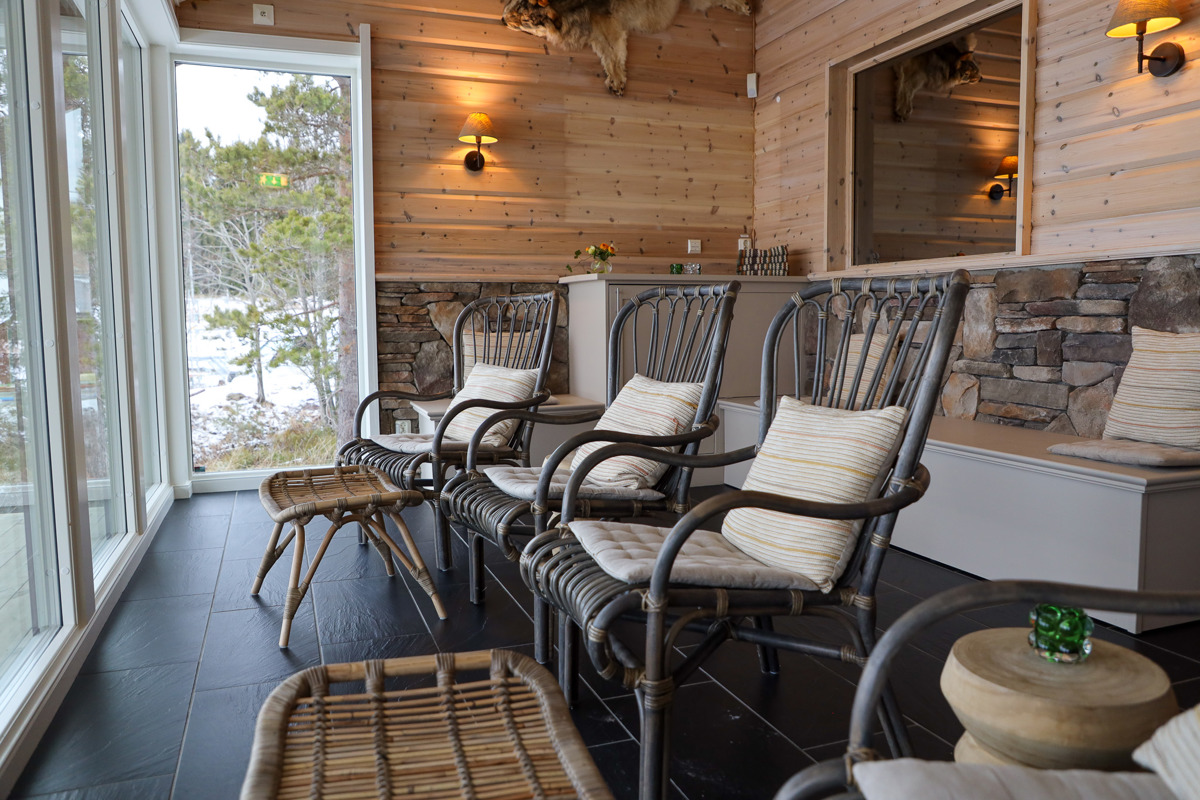 Avslappningsrum med rottingfåtöljer, kuddar och stora fönster mot vinterlandskap.