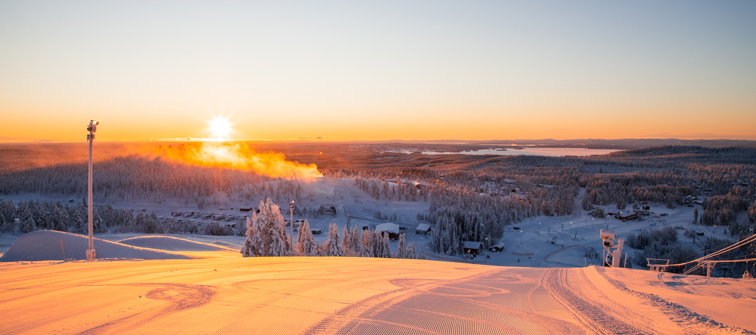 Soluppgång över en nypreparerad skidbacke med utsikt över ett snötäckt landskap och sjöar i fjärran.