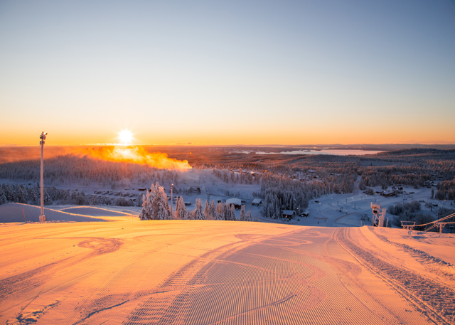 Sunrise over a freshly groomed ski slope with a view of a snow-covered landscape and distant lakes.