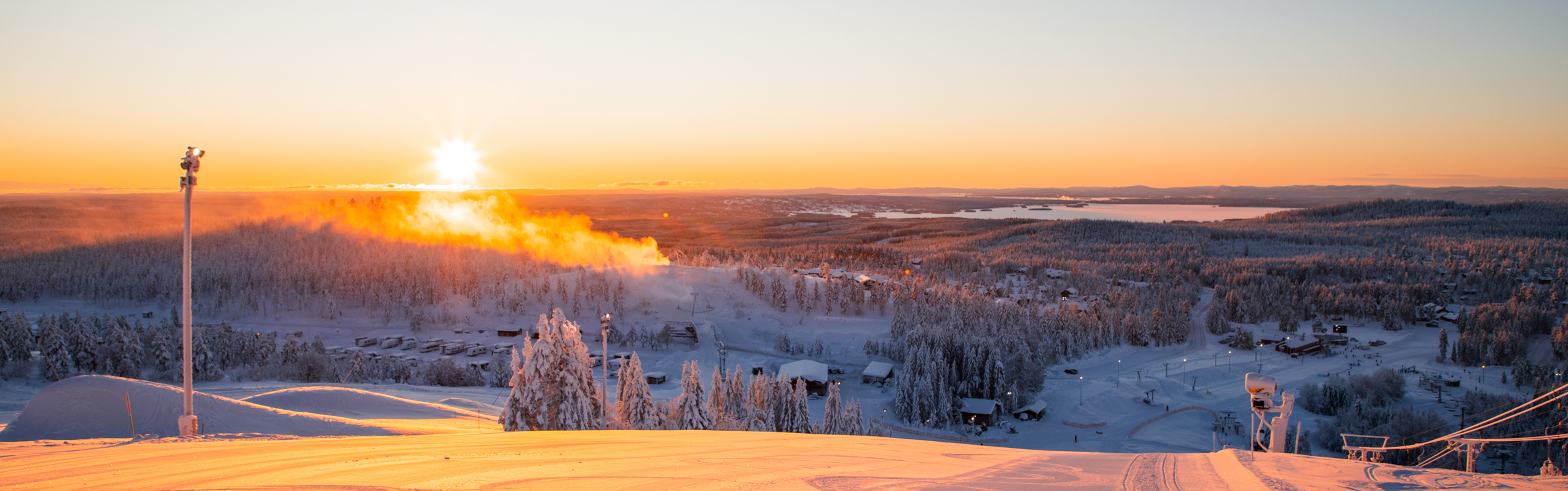 Soluppgång över en nypreparerad skidbacke med utsikt över ett snötäckt landskap och sjöar i fjärran.
