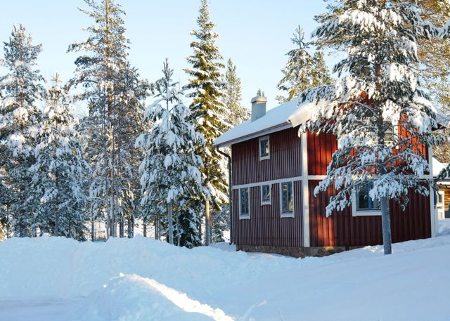 Röd stuga i Orsa Grönklitt omgiven av snötyngda granar i vintersolens sken.