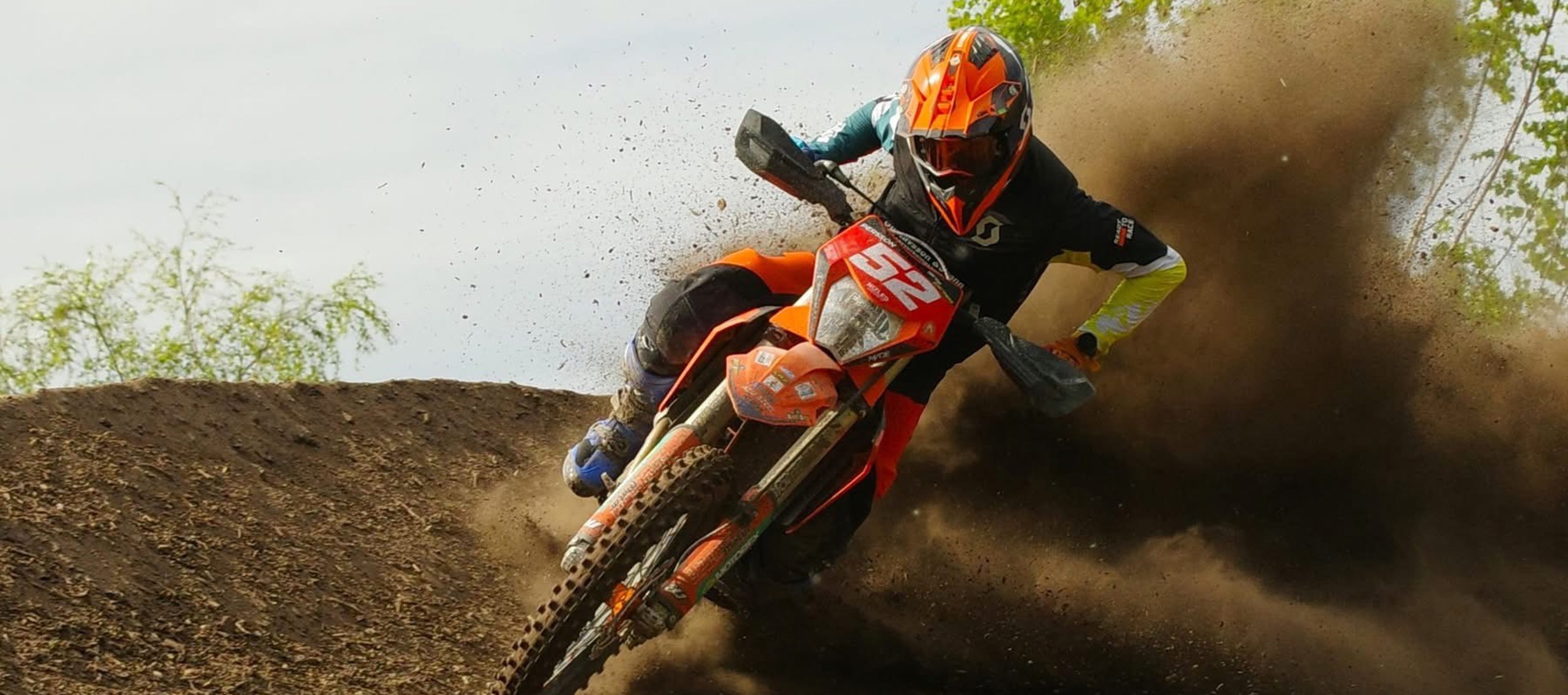 Motocrossförare i orange hjälm kör i hög fart genom en kurva, med jord som yr upp bakom motorcykeln.