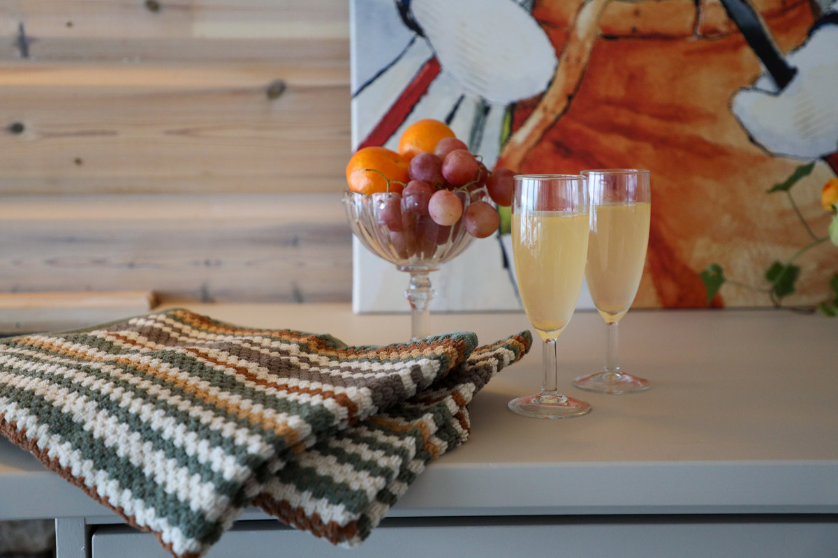 Two glasses of juice, a crocheted blanket, and a bowl of fruit on a table in front of a colorful painting.