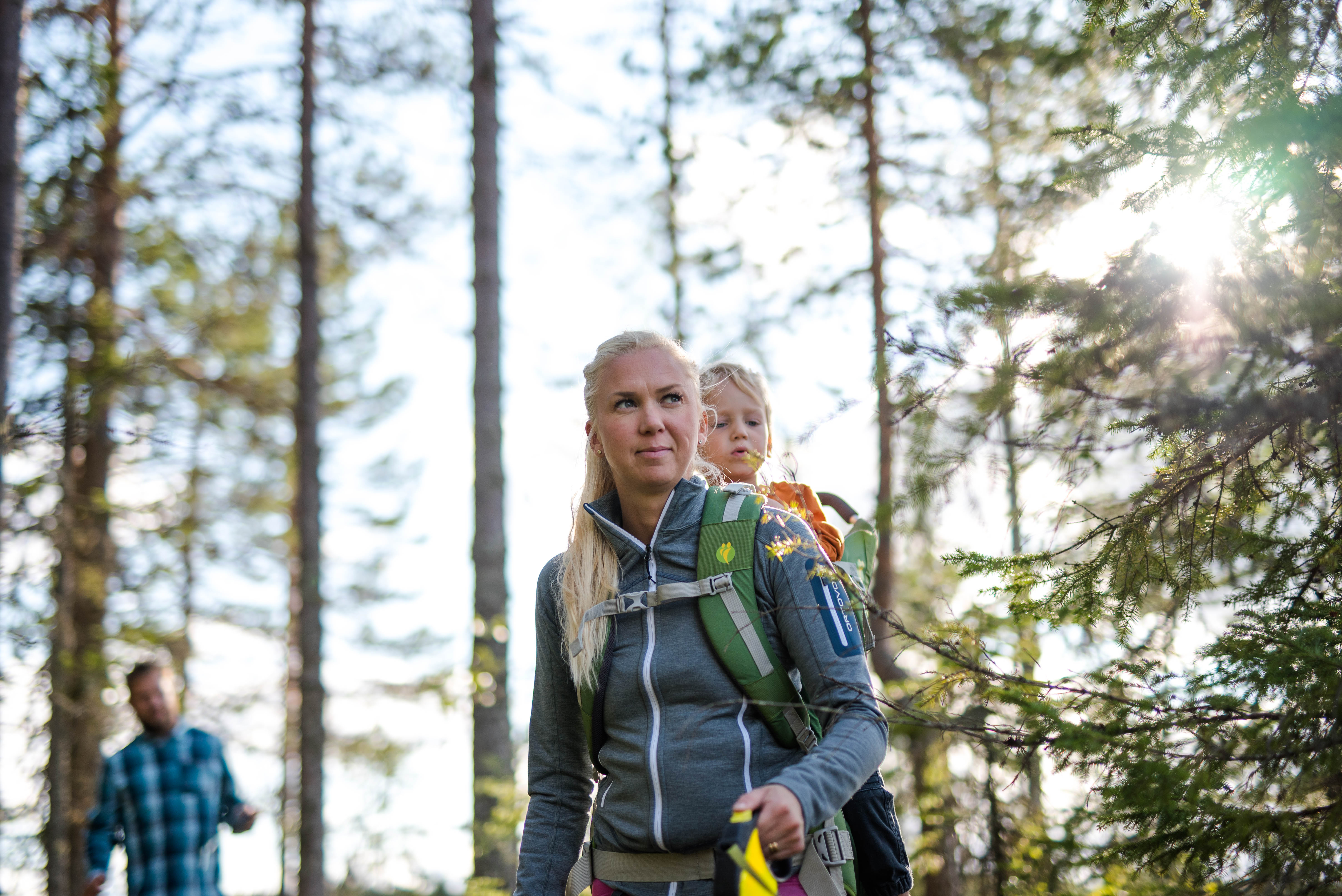 Kvinna vandrar i skogen med ett barn i bärstol på ryggen, med en man i bakgrunden.