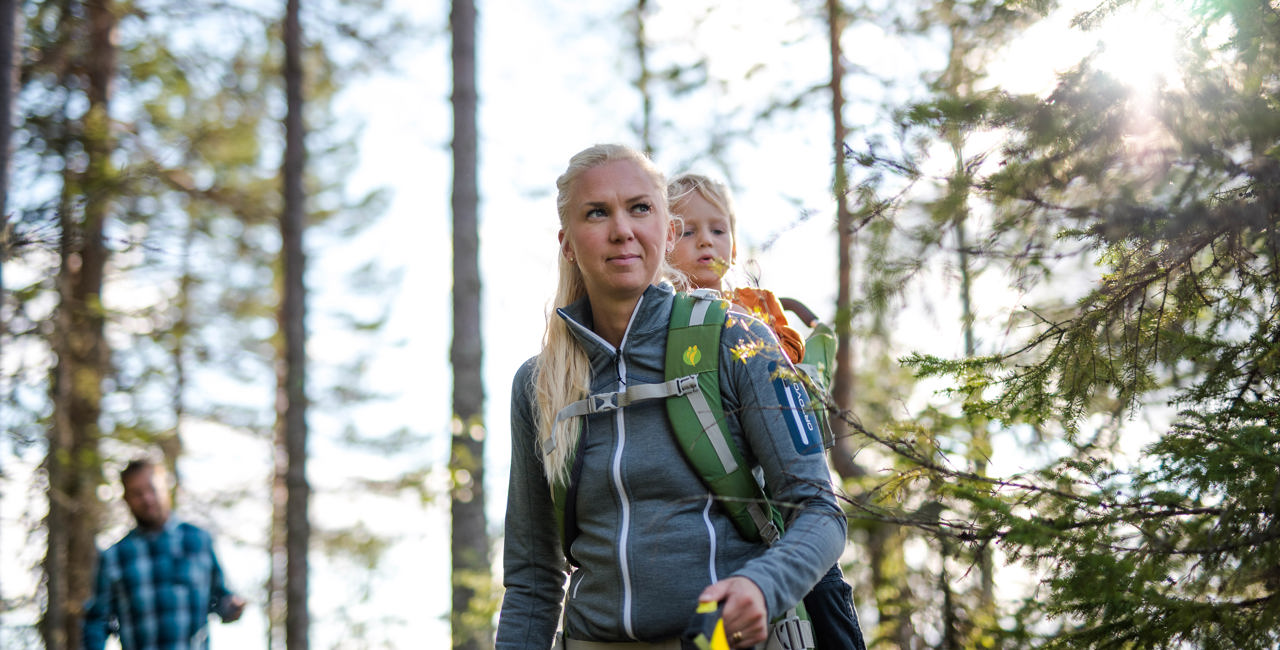 Kvinna vandrar i skogen med ett barn i bärstol på ryggen, med en man i bakgrunden.