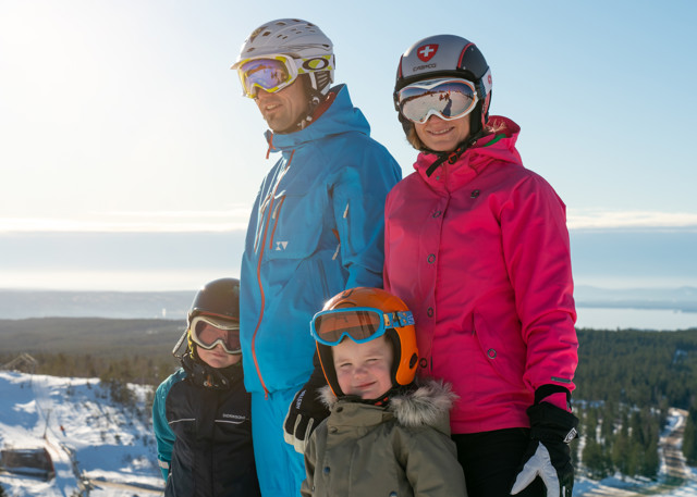 En familj i skidkläder står tillsammans på en snötäckt höjd med utsikt över landskapet.