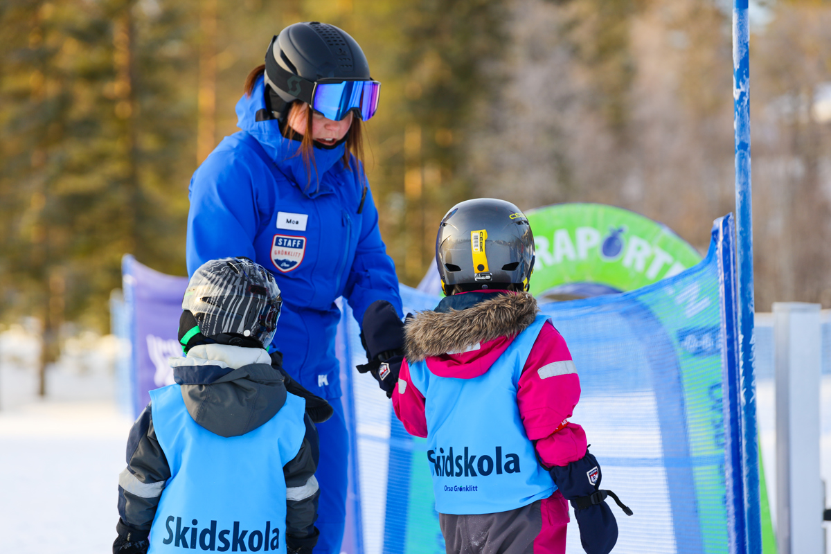 Ski instructor in a blue jacket helping two children at the ski school in Orsa Grönklitt. The children wear blue vests labeled “Skidskola”.