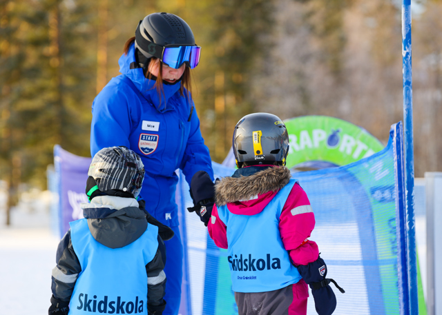 Ski instructor in a blue jacket helping two children at the ski school in Orsa Grönklitt. The children wear blue vests labeled “Skidskola”.