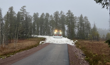Pistmaskin sprider snö på en väg omgiven av skog och höstfärgad mark i dimmigt väder i Orsa Grönklitt.