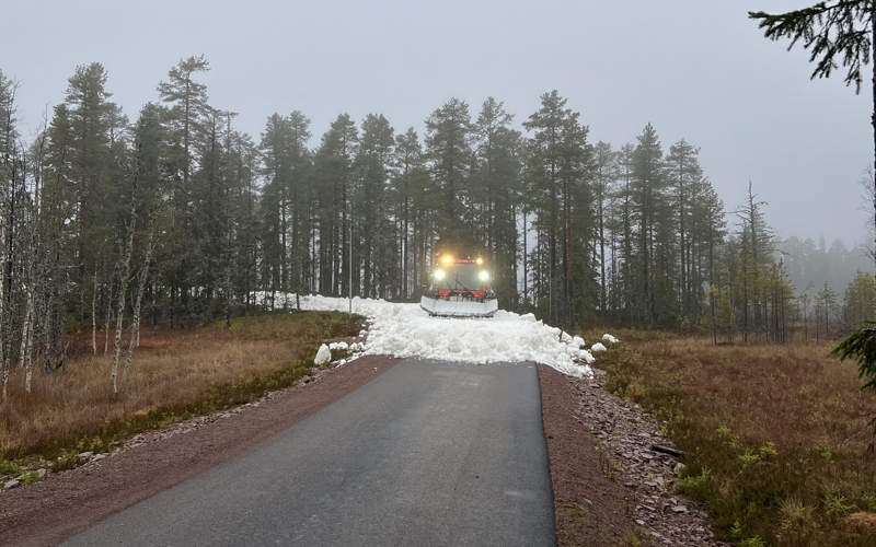 Pistmaskin sprider snö på en väg omgiven av skog och höstfärgad mark i dimmigt väder i Orsa Grönklitt.