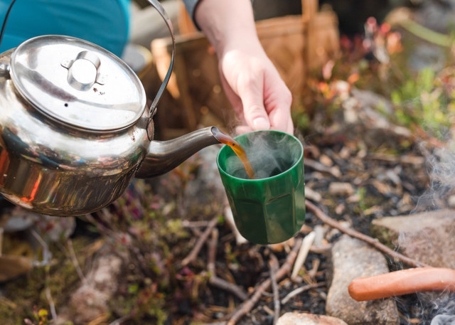 Kaffe hälls från kittel i en mugg vid en eldstad utomhus, med grillkorv i förgrunden.