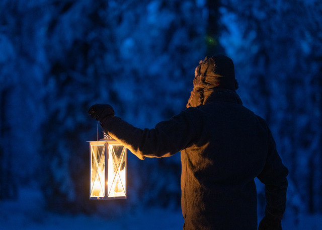 A person walks through a snow-covered forest at dusk, lighting the way with a glowing lantern