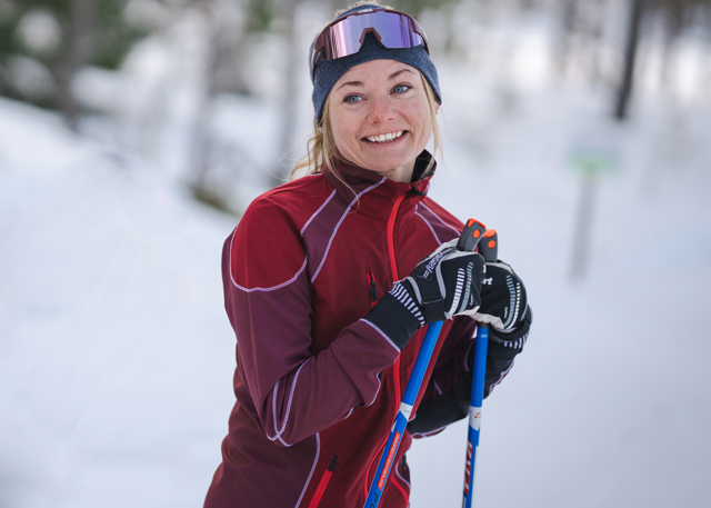 En leende kvinna i vinröd träningsjacka och pannband står med längdskidstavar i snön, omgiven av vinterlandskap.