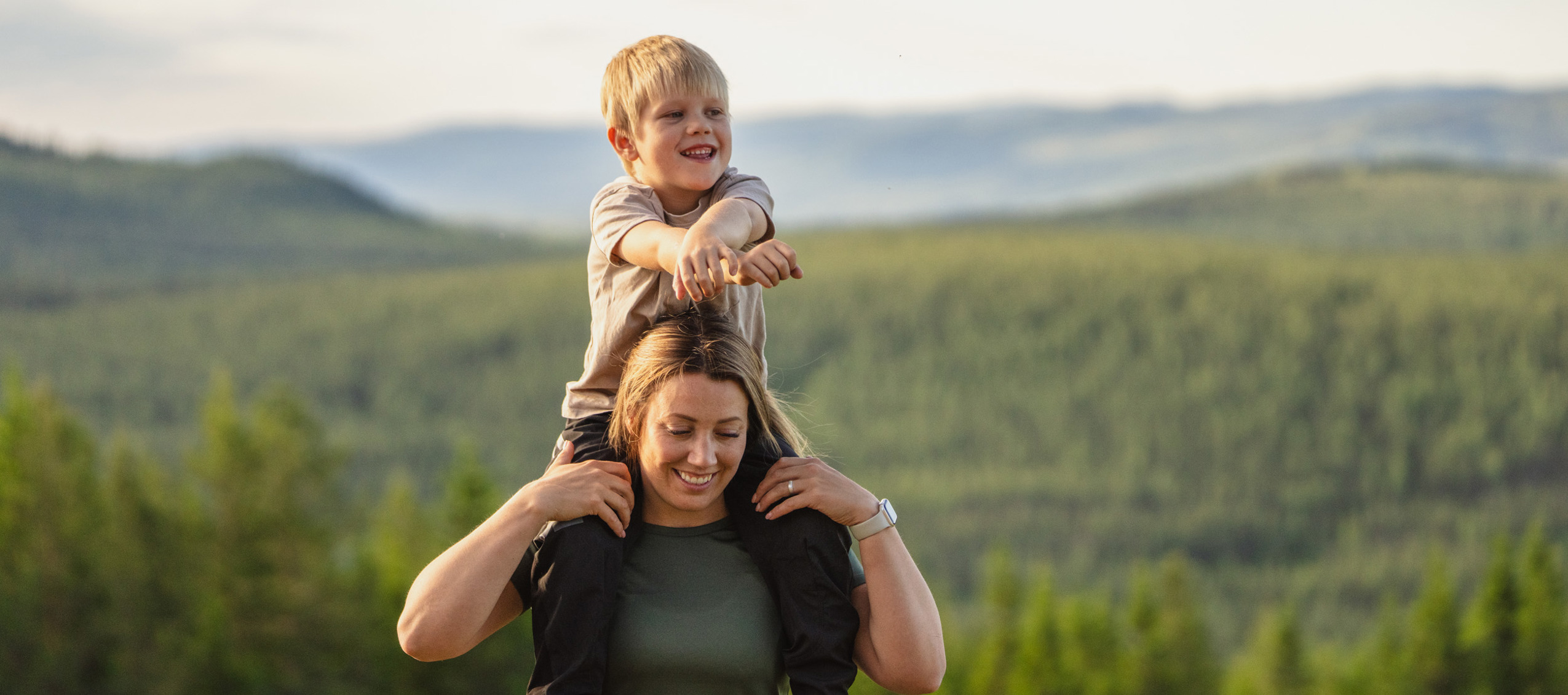 Leende barn på en kvinnas axlar med utsikt över gröna skogar och berg i Orsa Grönklitt.