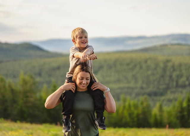 Leende barn på en kvinnas axlar med utsikt över gröna skogar och berg i Orsa Grönklitt.