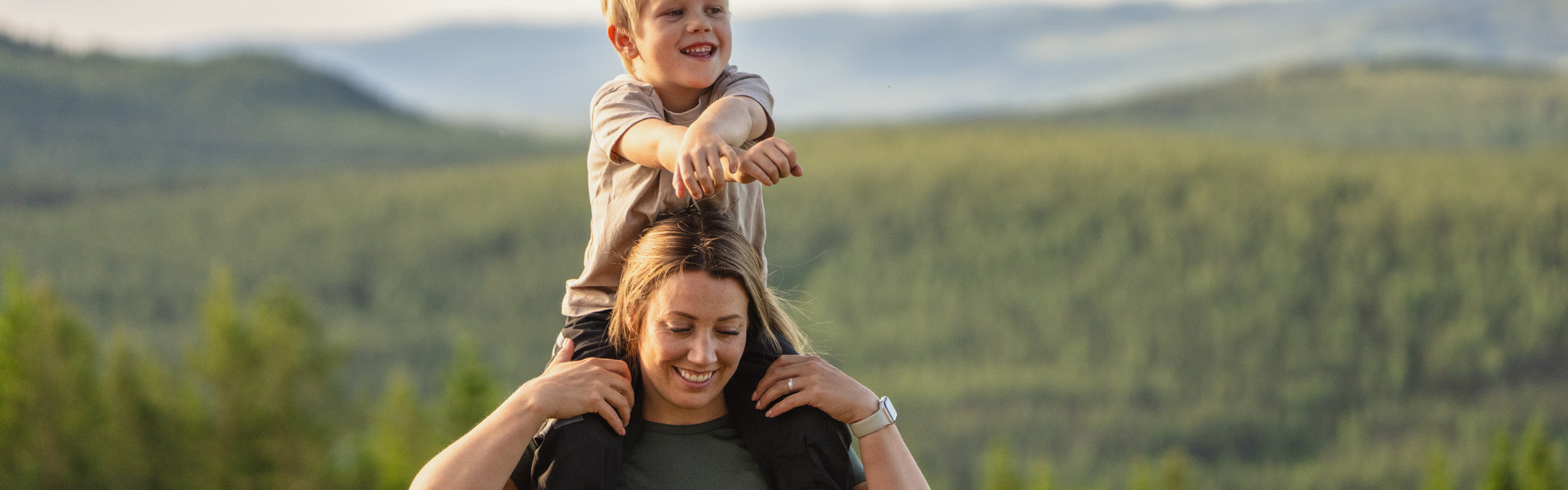 Smiling child on a woman's shoulders with a view of green forests and mountains at Orsa Grönklitt.