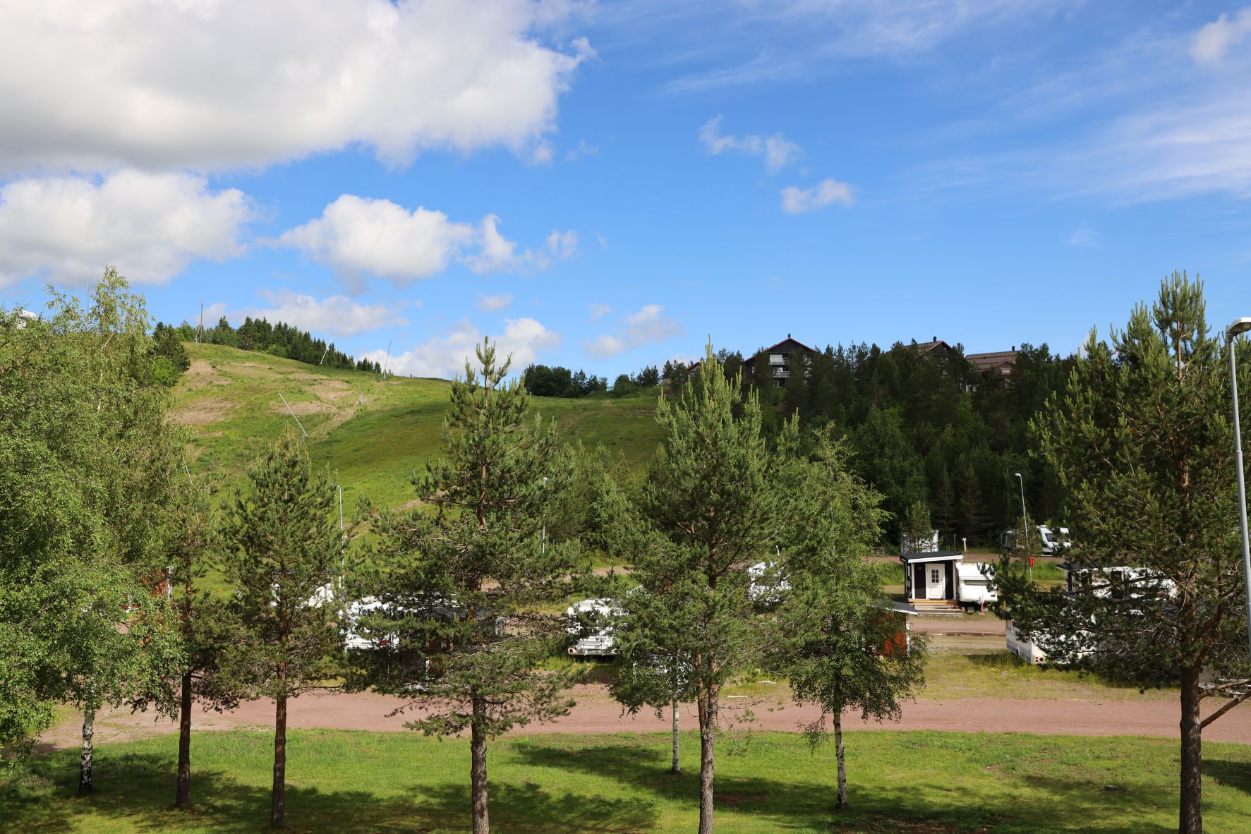 Sommervy over en grøn skibakke med blå himmel, hvide skyer og frodige træer i forgrunden.