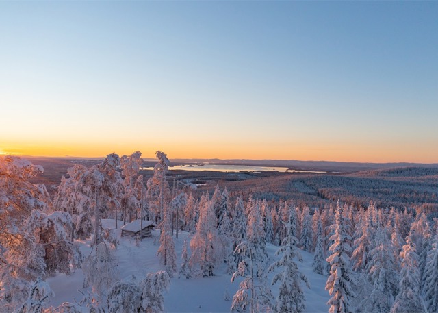 Soluppgång över ett vinterlandskap med snötäckta trädtoppar och utsikt över fjäll, skogar och sjöar i horisonten.