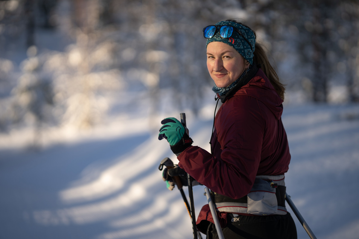 A woman in a red jacket and headband cross-country skiing in a sunny winter landscape. She smiles at the camera