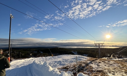 Snötäckt skidbacke i solnedgång med liftstolpar, liftvajrar och utsikt över skog och landskap i fjärran.