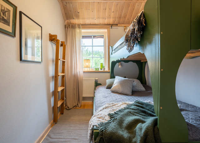 Ett mysigt sovrum med våningssäng i grönt trä, bäddad med filtar och kuddar. Fönstret släpper in dagsljus och rummet har träpanel i taket samt en stege som leder till överslafen.