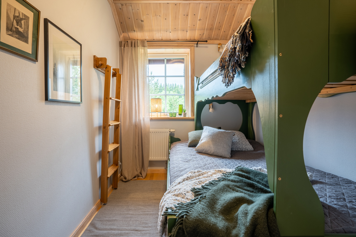 Ett mysigt sovrum med våningssäng i grönt trä, bäddad med filtar och kuddar. Fönstret släpper in dagsljus och rummet har träpanel i taket samt en stege som leder till överslafen.