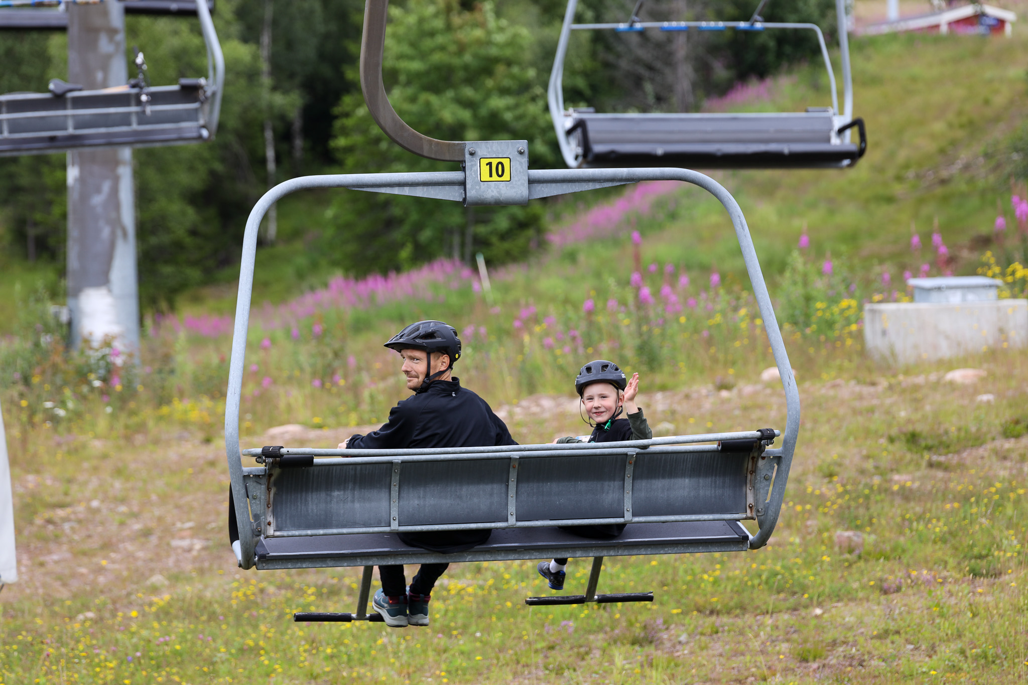 En voksen og et barn med cykelhjelme sidder i en stolelift på vej op ad en grøn skråning med blomstrende pileurt i baggrunden.