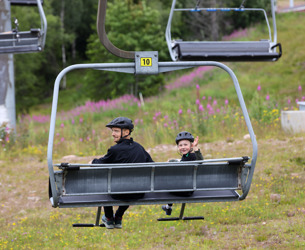 En vuxen och ett barn med cykelhjälmar sitter i en stollift på väg uppför en grön sluttning med blommande rallarrosor i bakgrunden.