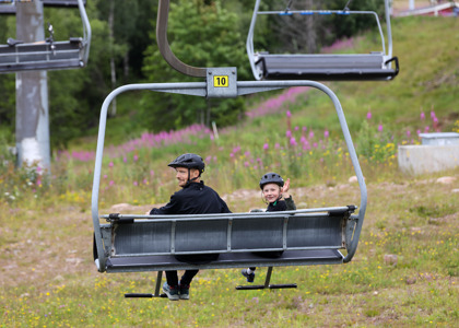 En vuxen och ett barn med cykelhjälmar sitter i en stollift på väg uppför en grön sluttning med blommande rallarrosor i bakgrunden.