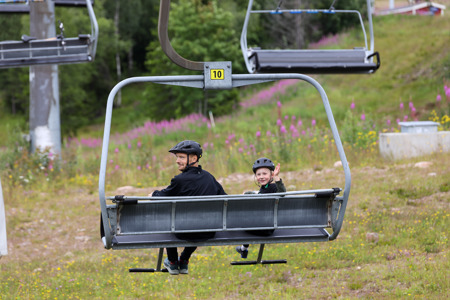 En vuxen och ett barn med cykelhjälmar sitter i en stollift på väg uppför en grön sluttning med blommande rallarrosor i bakgrunden.